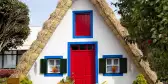Small rural house with a triangular thatched roof, a red front door and small windows with shutters.