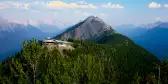 Large tree filled Sulphur Mountain with a building on the top of it, with snowy mountains in the background