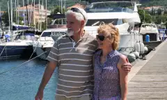 A male and female couple stood on the end of a jetty holding one another round the waist and arm with the sun on their faces.