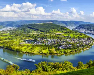 A wide shot of a bend in the Moselle River from it's surrounding hills under broad daylight, two river cruise ships can be seen creating gentle waves in the water.