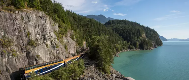 Aerial shot of dark blue and orange train surrounded by steep, rocky-mountains, tall pine trees and overlooking the ocean